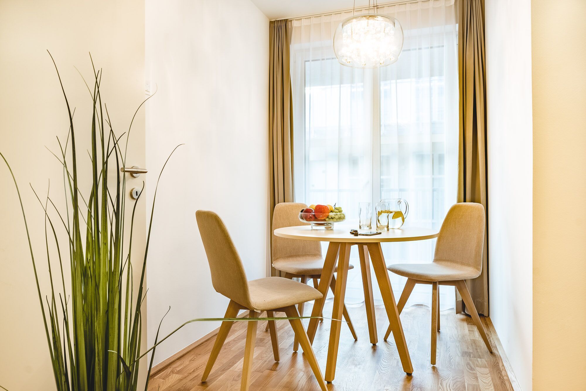 A dining area with a round table, four chairs, a chandelier, and a window with curtains.