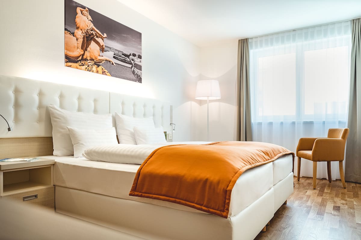 Quarto de hotel com cama, mesa de cabeceira, luminária, cadeira e uma grande janela com cortinas.