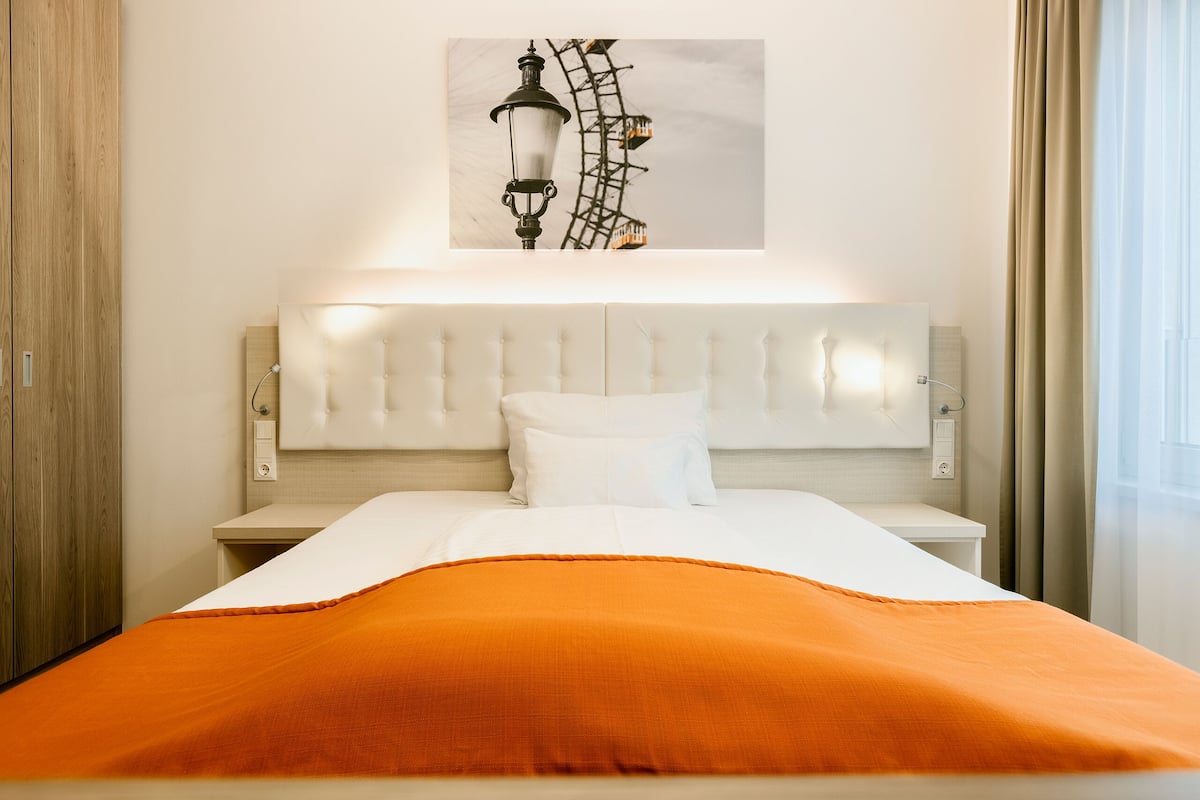 Uma cama bem arrumada com cabeceira branca e manta laranja, uma luminária fixada na parede e uma imagem de montanha-russa.