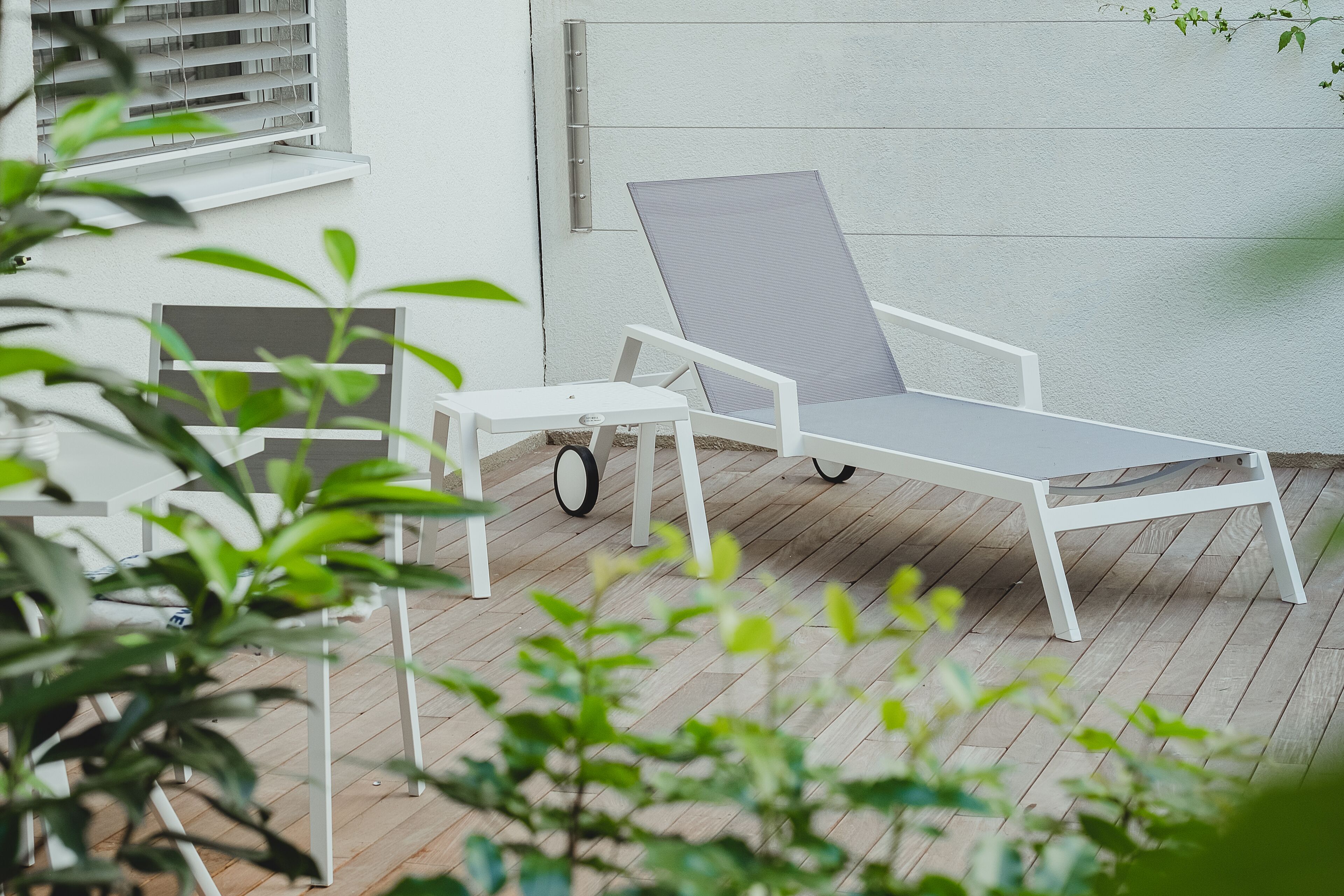 A patio with a white lounge chair and table, a window with blinds, and a white wall.