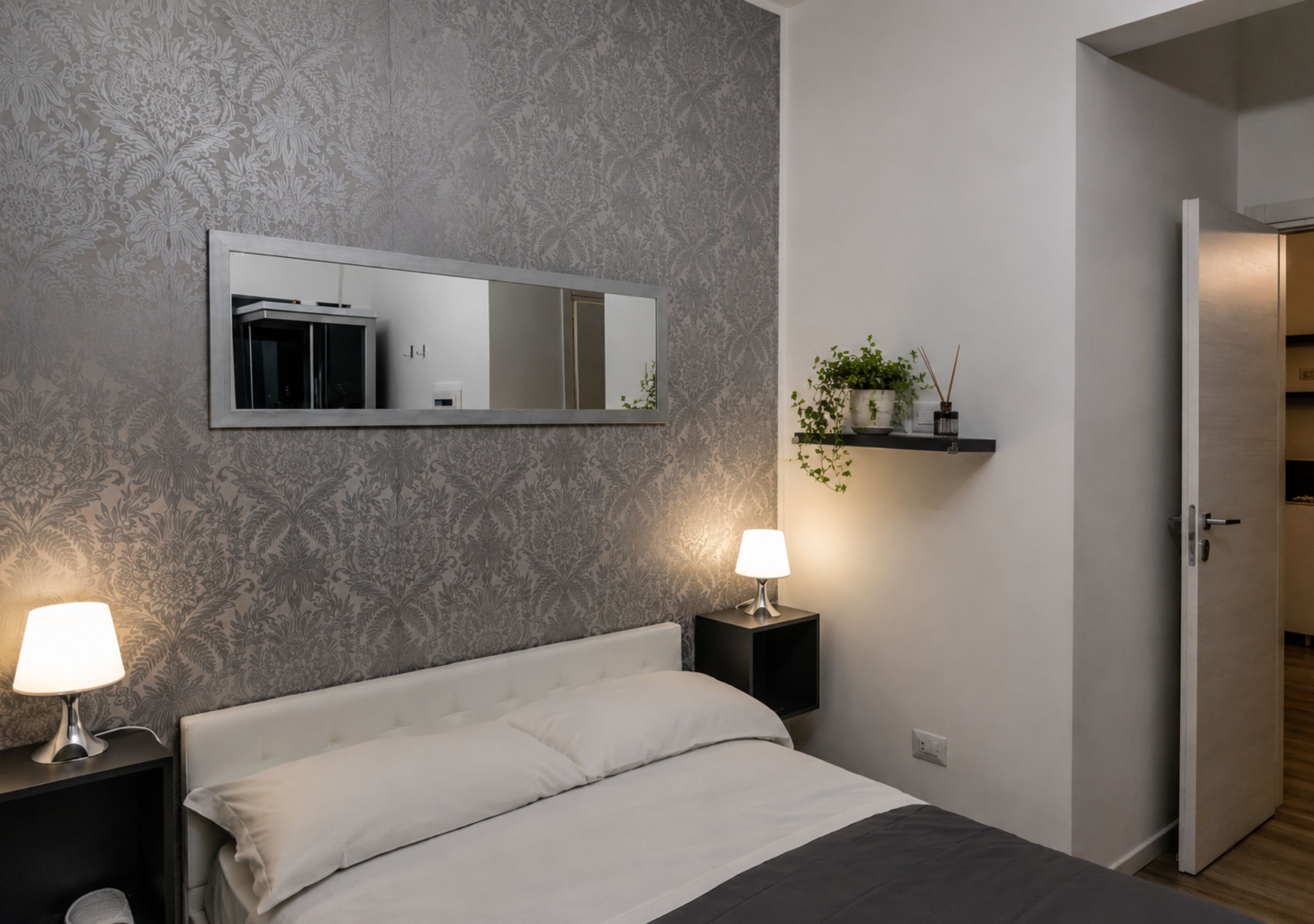 A bedroom with a bed, nightstands with lamps, a mirror, a wall-mounted shelf with plants, and a door.