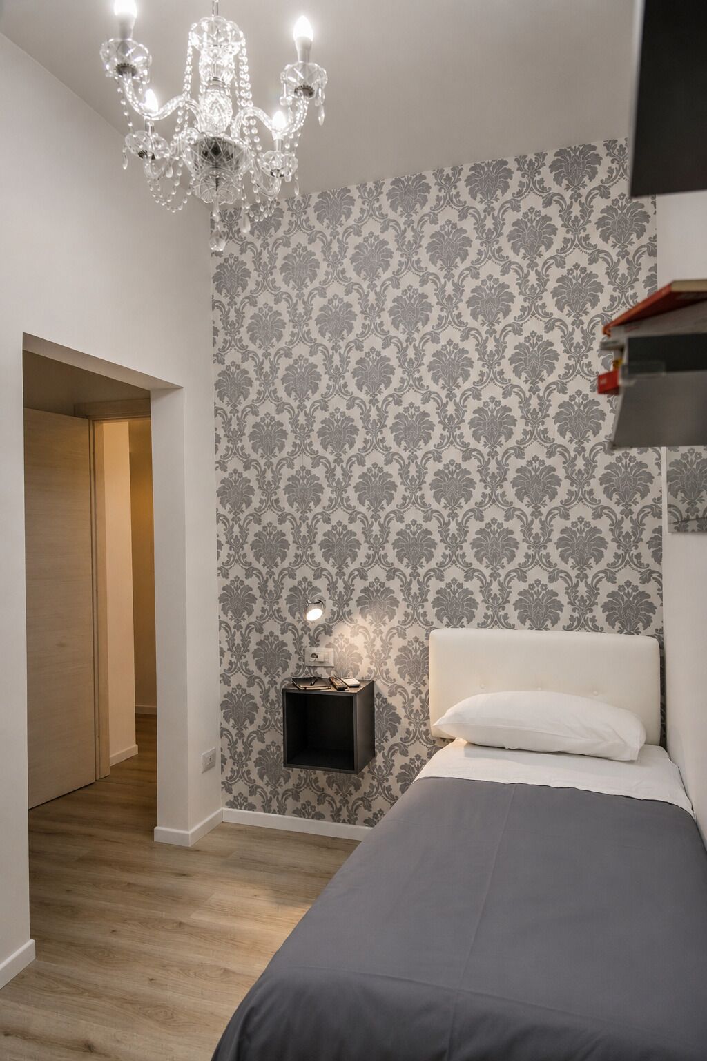 A bedroom with a bed, a nightstand with a lamp, a chandelier, and a patterned wallpaper.