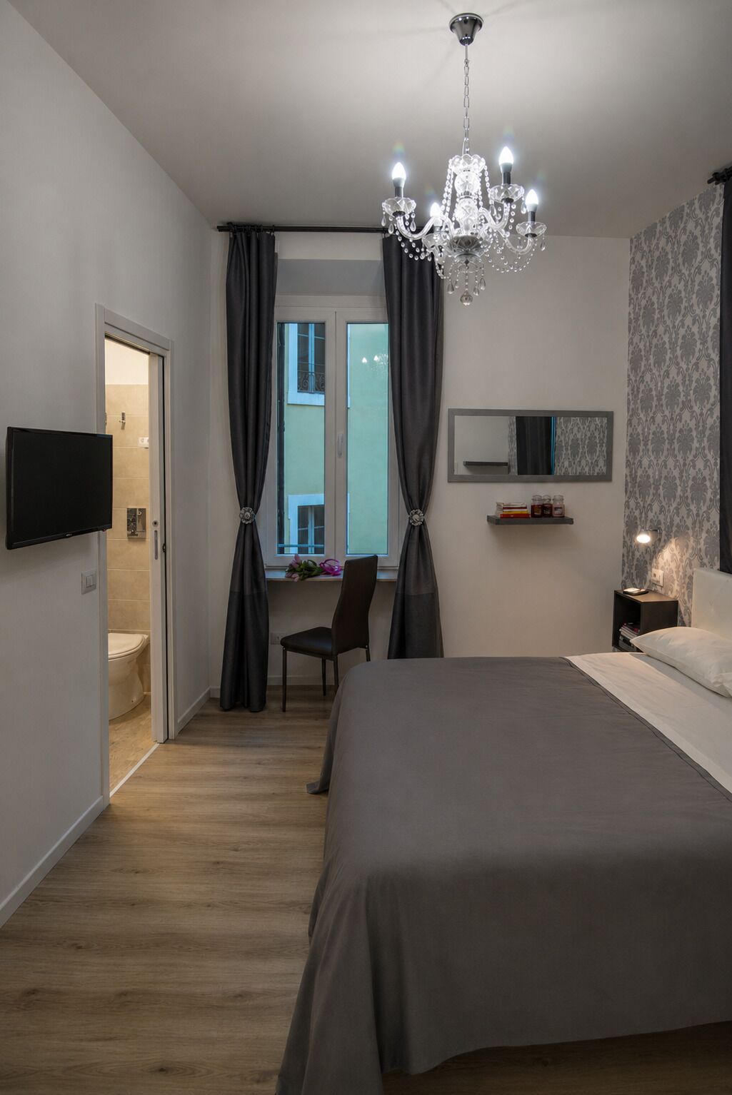 A bedroom with a bed, a TV, a desk, a chair, a window with curtains, a chandelier, and a nightstand.
