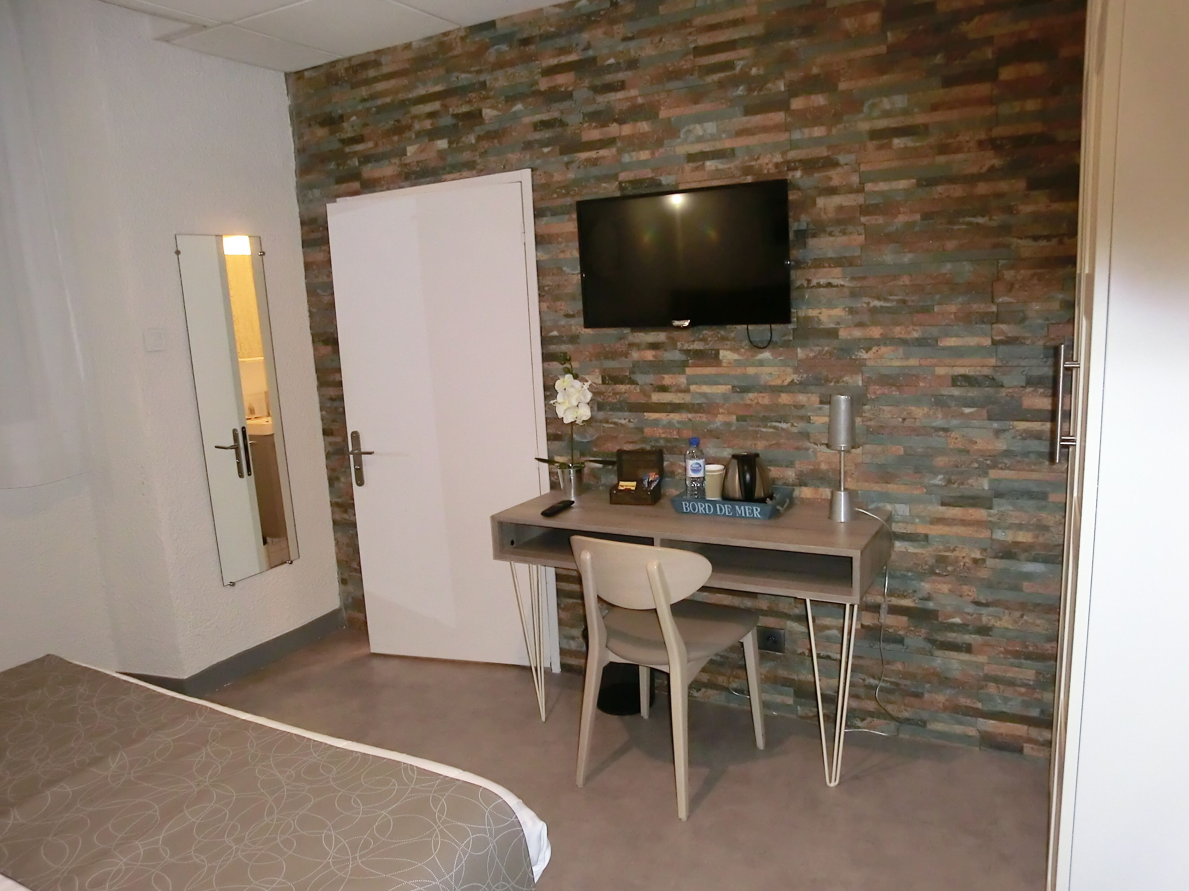 A hotel room with a brick-patterned wall, a flat-screen TV mounted above a desk, a white door, and a bed with a patterned bedspread.