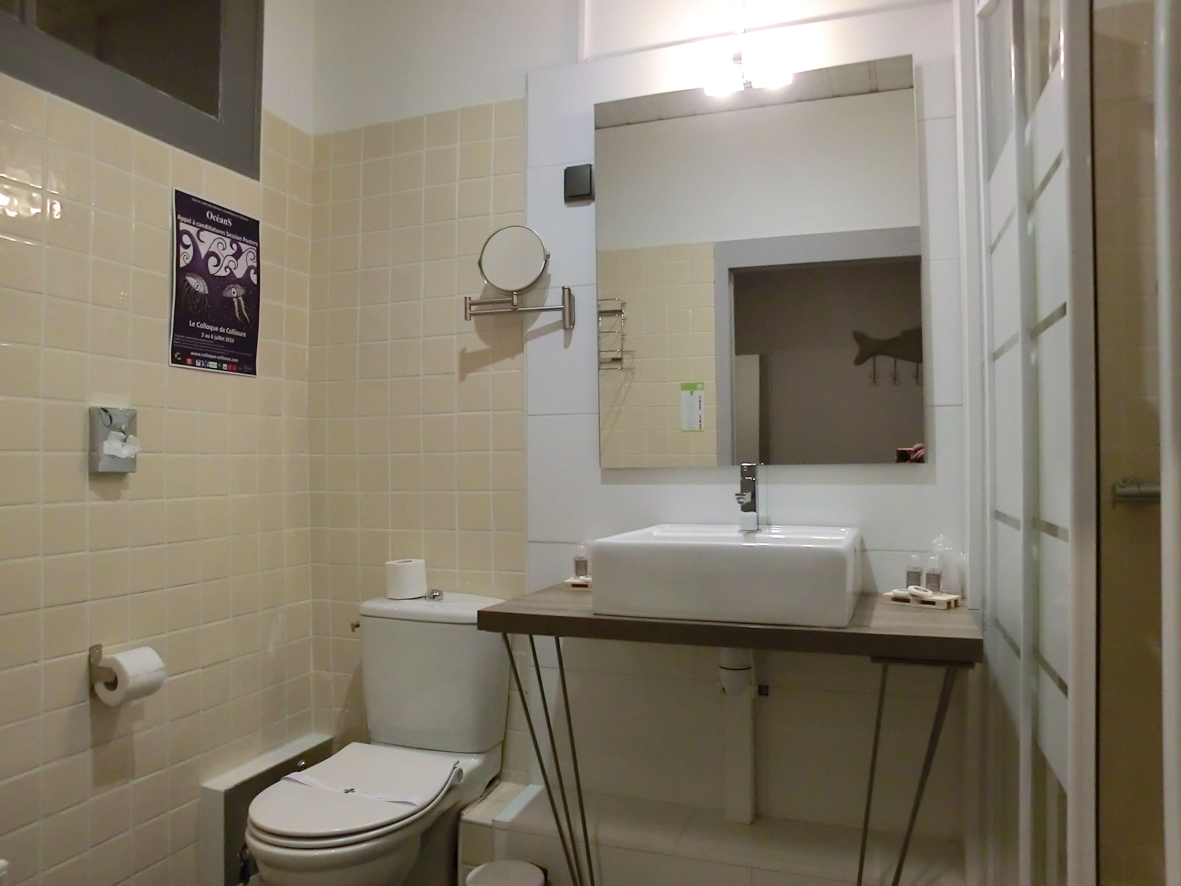 A bathroom with a toilet, sink, and mirror. There is a poster on the wall and a hand dryer.