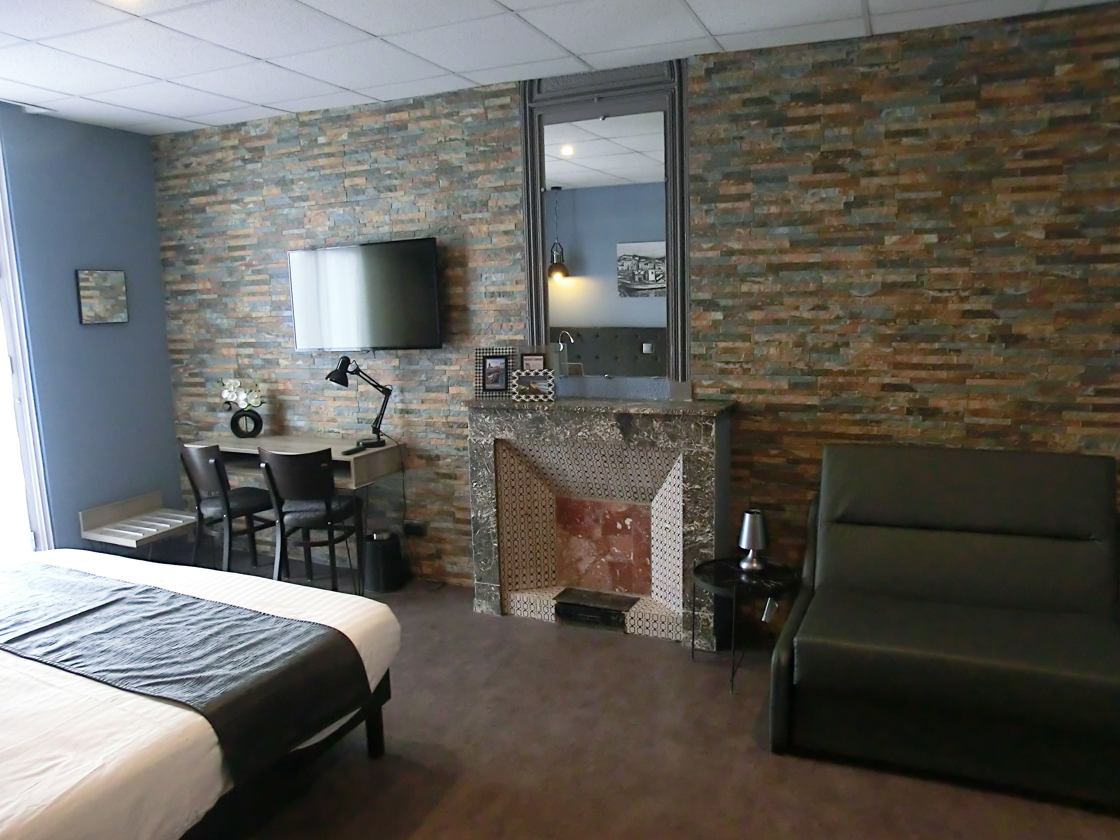 A hotel room with a brick wall, a flat-screen TV mounted above a fireplace, a bed with a dark headboard, a desk with a chair, and a large armchair.