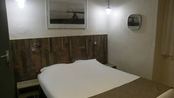 A hotel room with a wooden headboard, a bed with white bedding, a bedside table, a small mirror, and a framed picture on the wall.