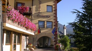 Ein Hotel namens "Hotel Corona" mit Balkonen, die mit Blumen geschmückt sind, einem klaren Himmel und Bergen im Hintergrund.