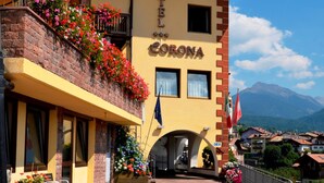 Ein Hotel namens "HOTEL CORONA" mit einer gelben Fassade, Blumenkästen am Balkon und Blick auf die Berge im Hintergrund.