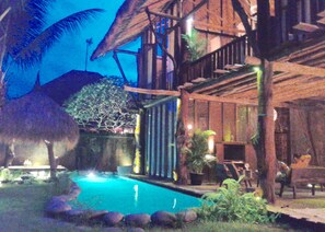 A pool area with a thatched-roof structure, a wooden deck, and surrounding greenery.