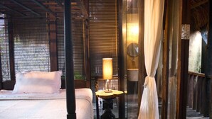 A four-poster bed with a white bedspread and pillows, a bedside table with a lamp, and a wooden floor. - Rustic Hut in Seminyak Bali (Seminyak)