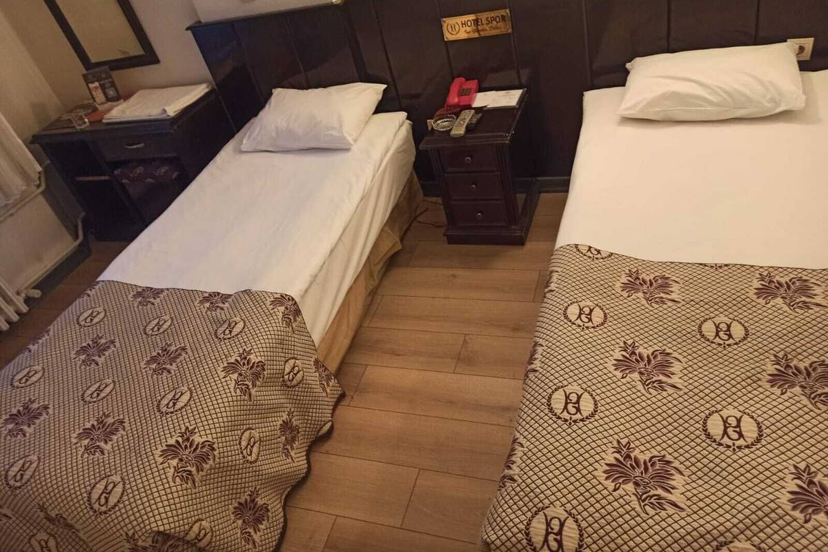 A hotel room with two beds, each with a white pillow and a patterned bedspread. There is a nightstand between the beds with a telephone and a small lamp. A sign on the wall reads "HOTEL SPORT."