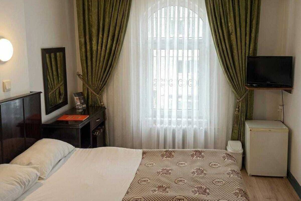 A hotel room with a bed, a TV, a small cabinet, and a window with green curtains.