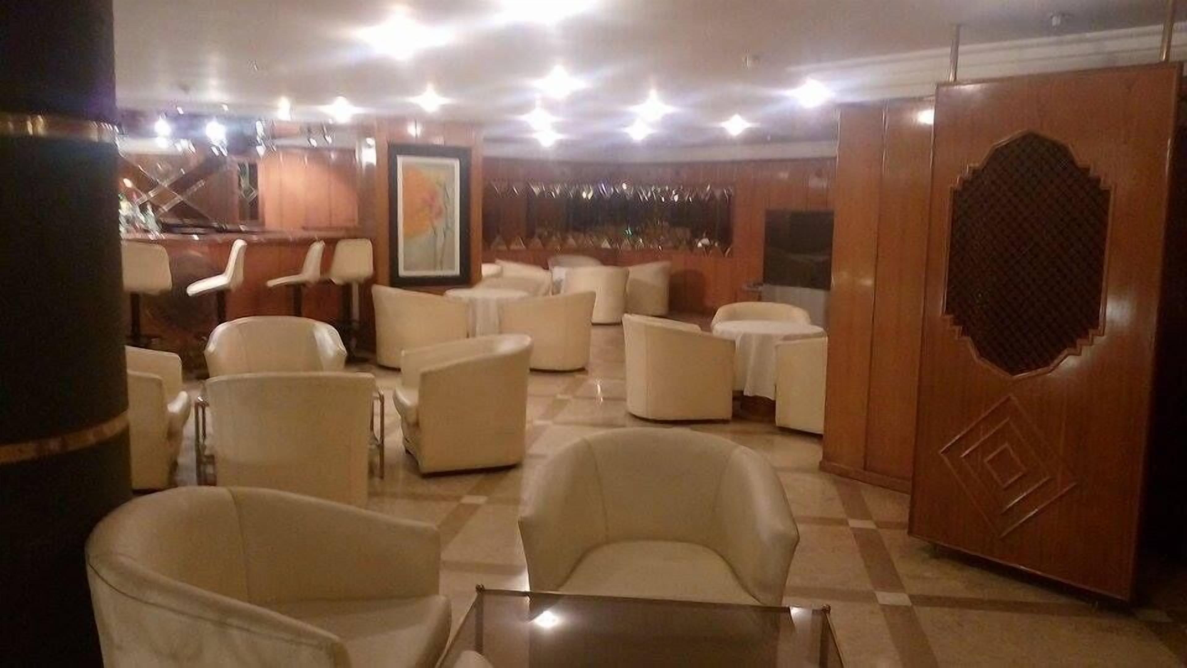 Un hall de hotel con varias zonas de estar, incluyendo sofás y sillones, organizados alrededor de una mesa de centro. Se observa una puerta de madera grande con un diseño decorativo en el lado derecho.