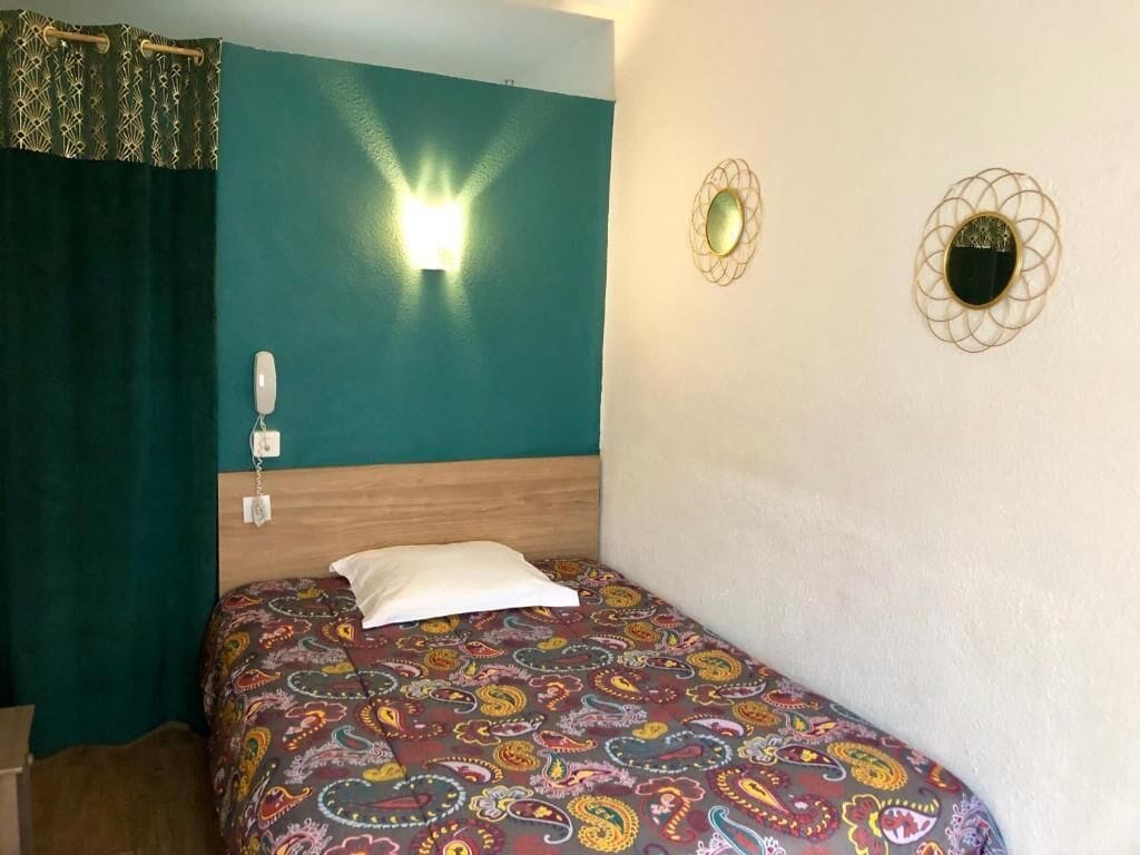 A single bed with a patterned bedspread, a wooden headboard, a wall-mounted light, and two wall sconces.