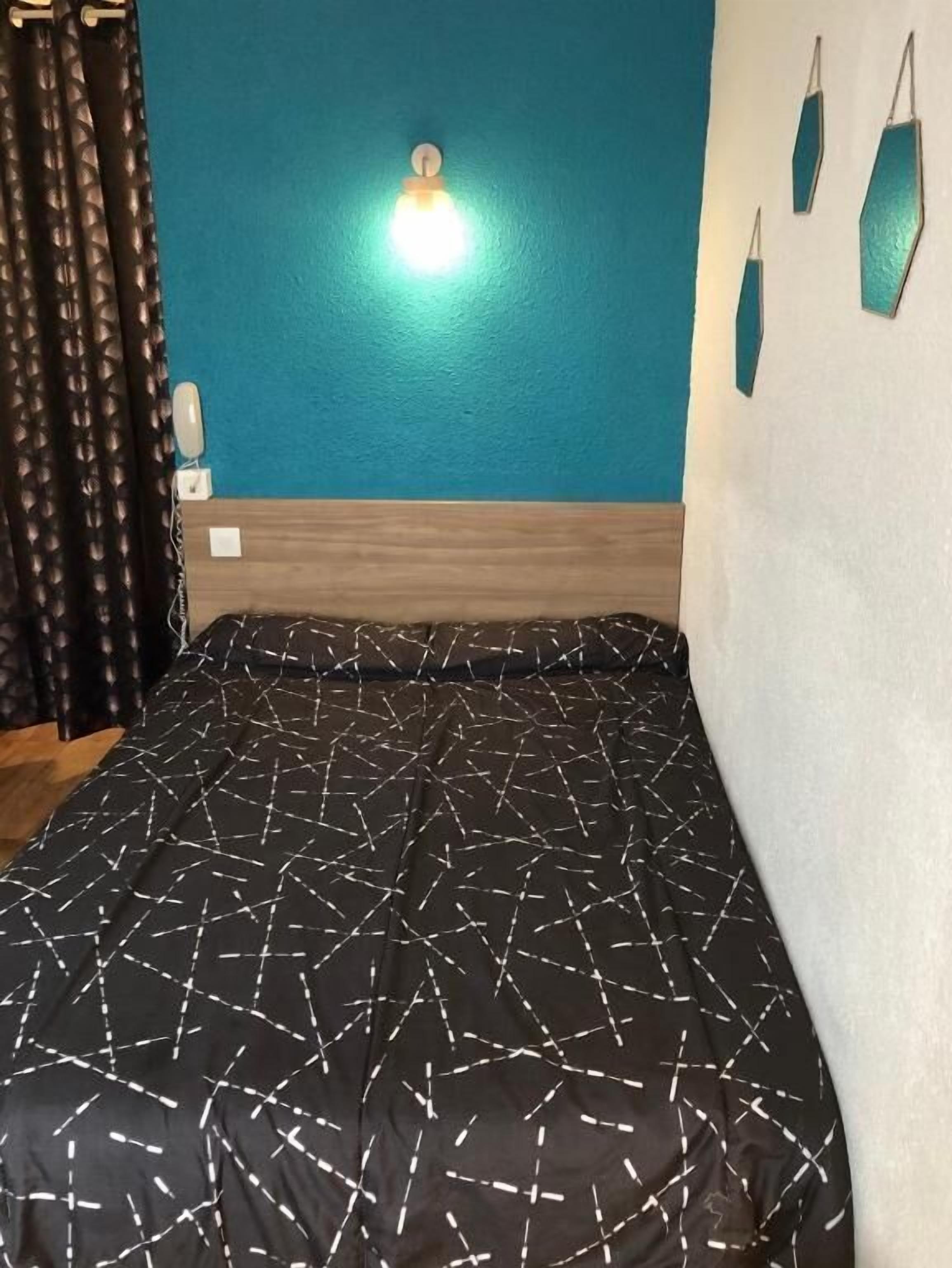 A bed with a geometric patterned bedspread, a teal wall with a light fixture, and three teal wall hangings.