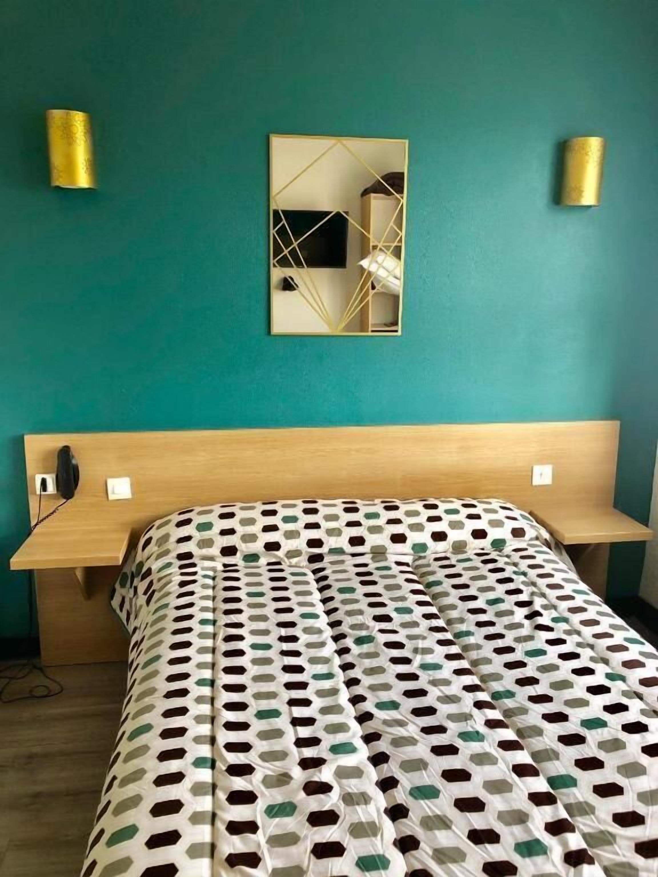 A bedroom with a teal wall, a wooden headboard, a patterned bedspread, and two wall-mounted lamps.
