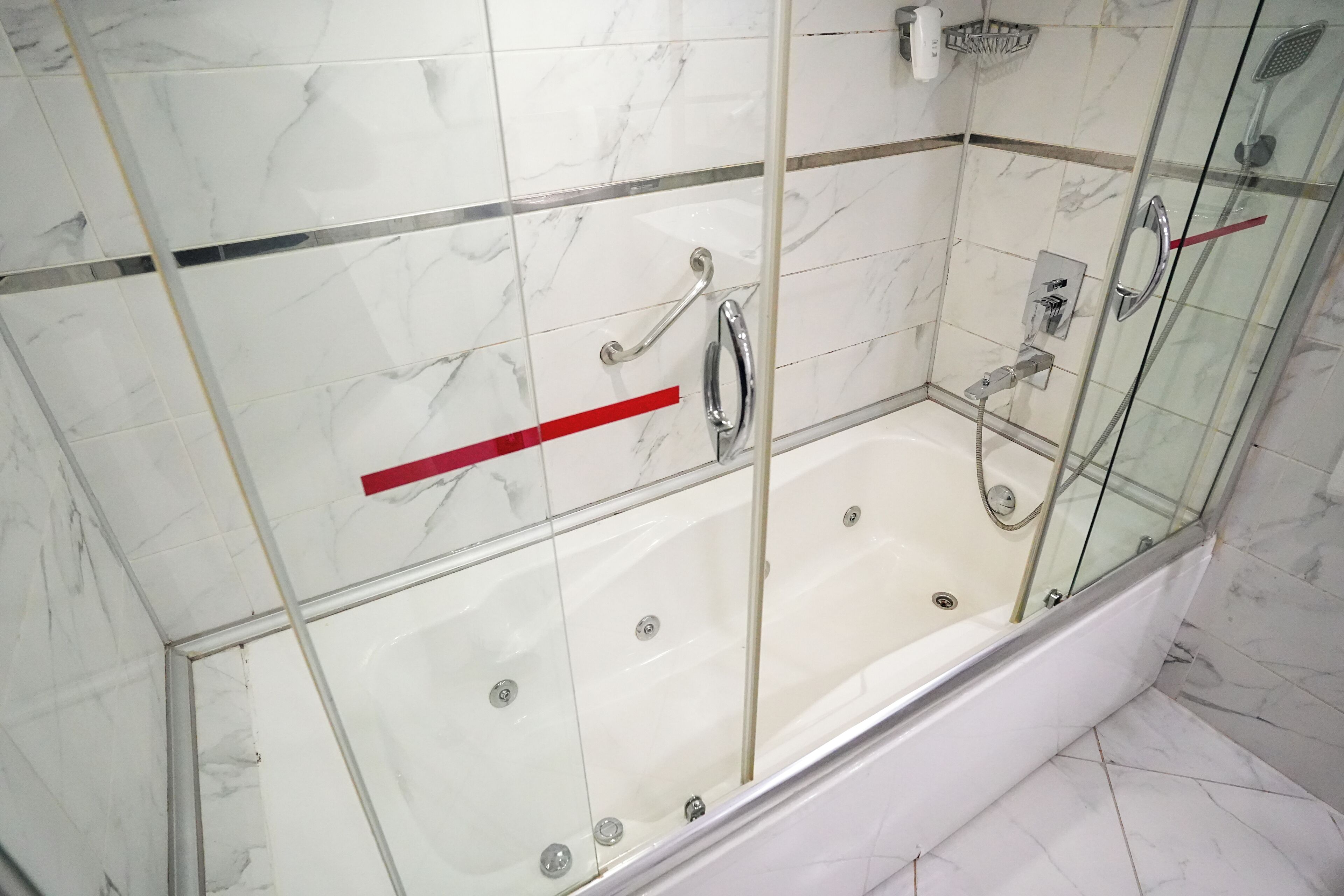 Un baño moderno con ducha envuelta en vidrio, azulejos de mármol y un pasamanos rojo.
