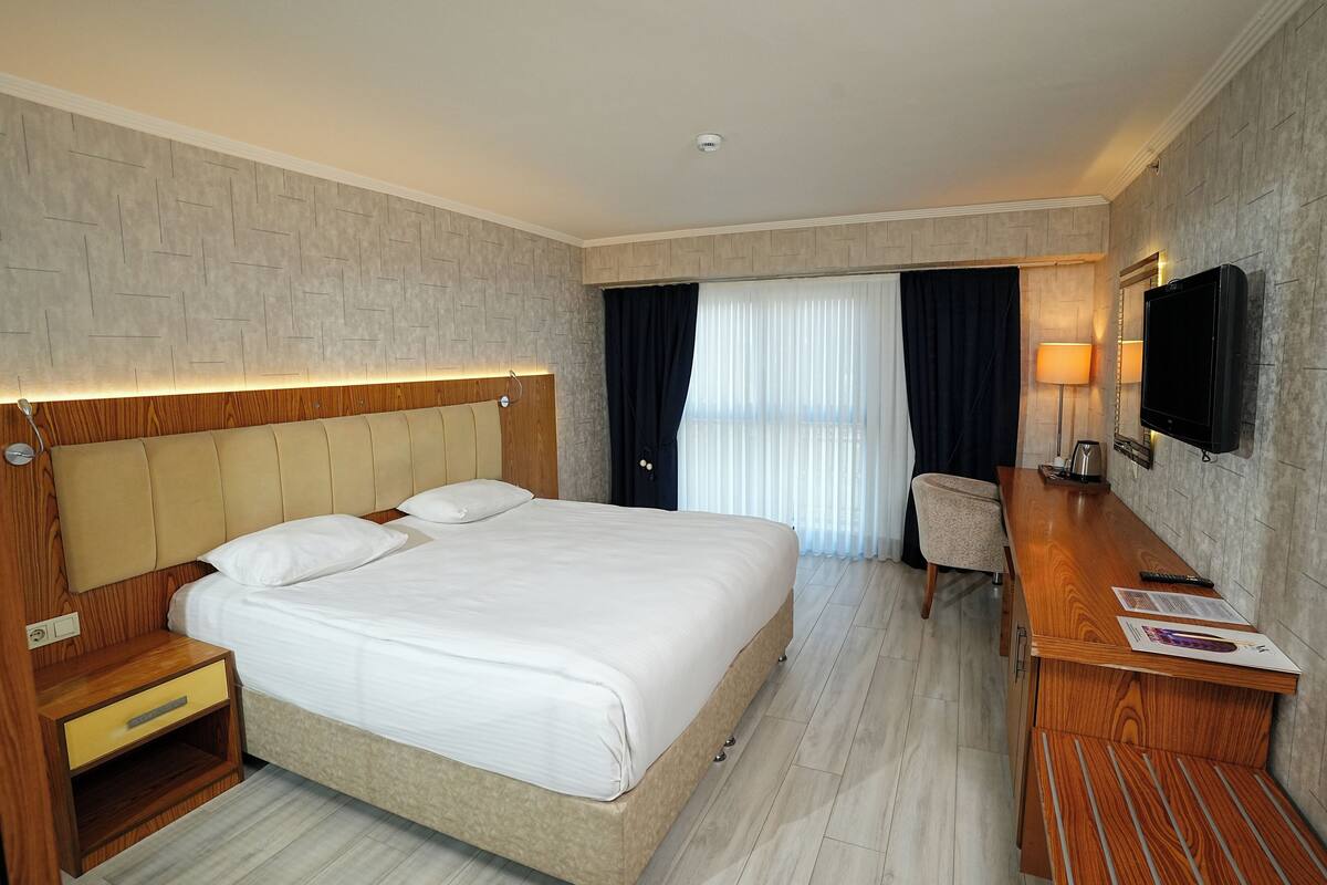 A hotel room with a large bed, bedside tables, a desk, a chair, a TV, and a window with curtains.
