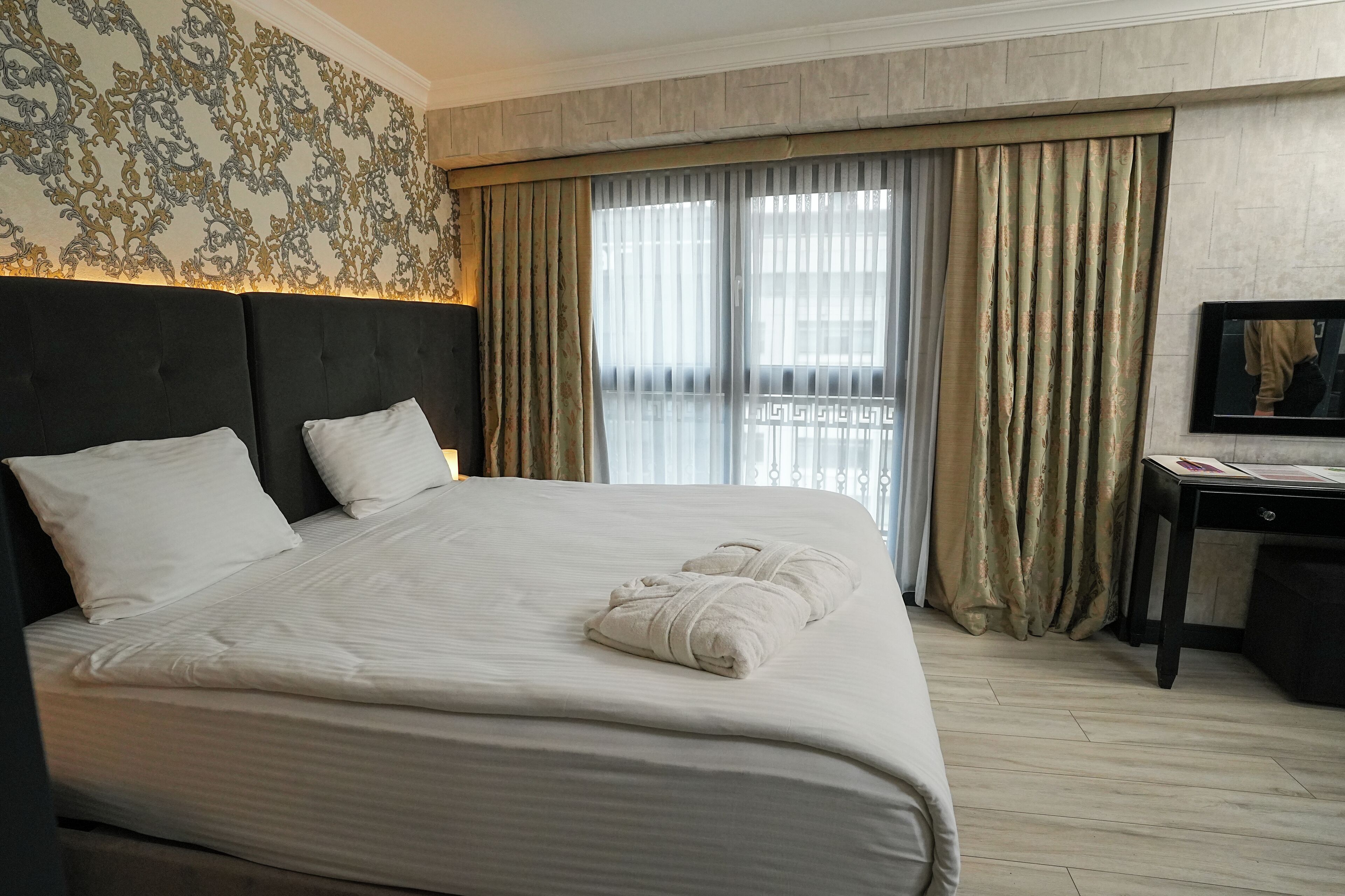 A neatly made bed with white linens and two pillows, a dark headboard, and a large window with sheer curtains.