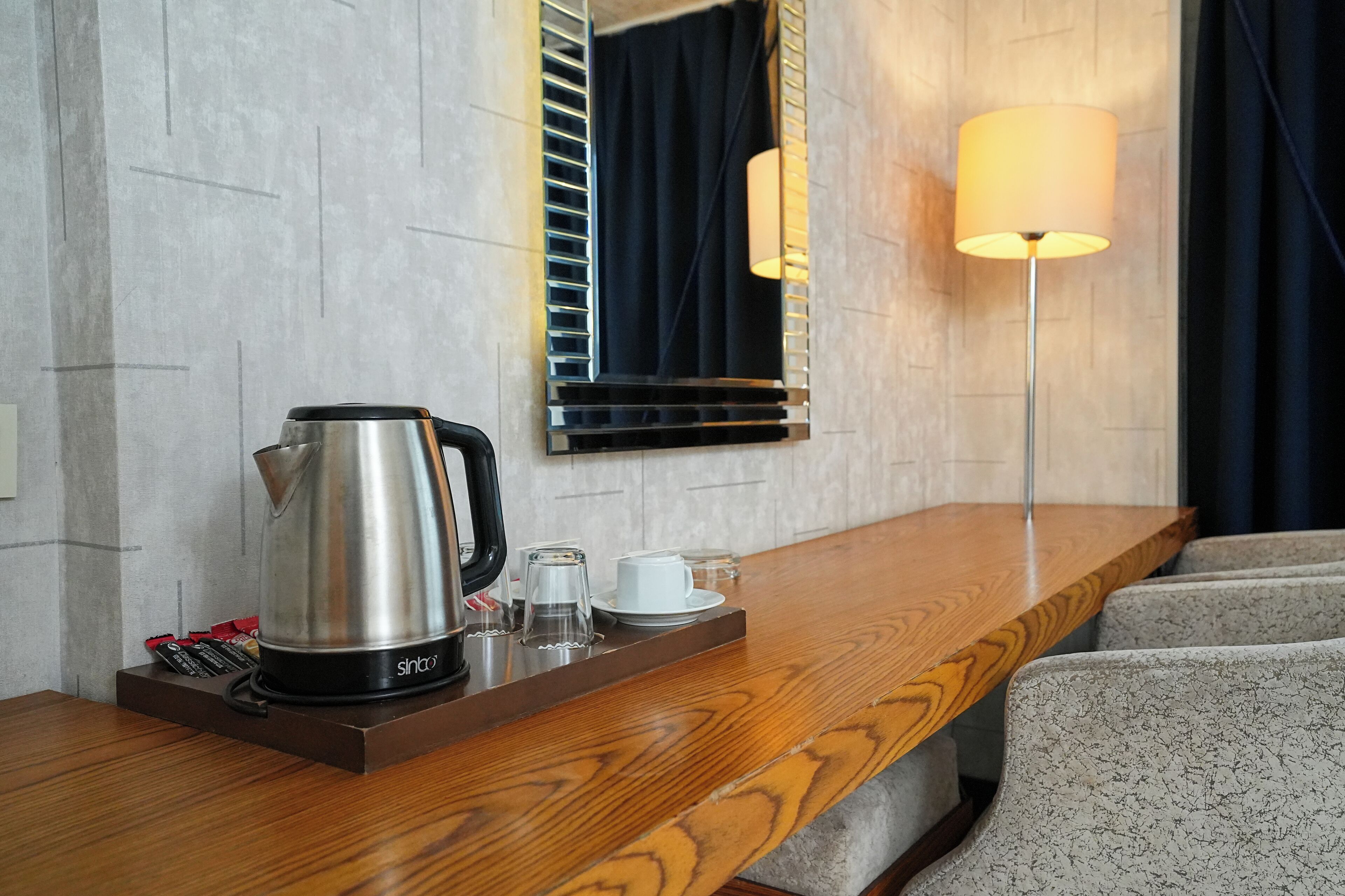 A hotel room with a wooden desk, a stainless steel coffee maker, a cup, and a glass.