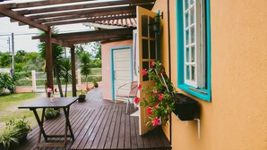 Une terrasse en bois avec une table et des chaises, une porte et une fenêtre agrémentée d’un pot de fleurs suspendu.