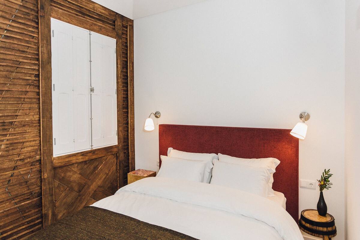 Un dormitorio con paneles de madera, cabecera roja, ropa de cama blanca y una mesita de noche con un jarrón.