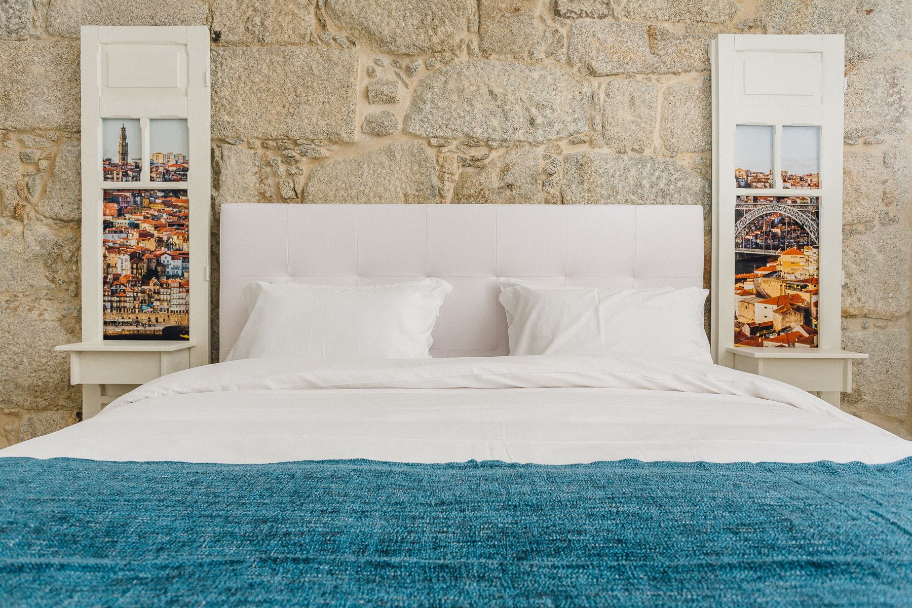 Un lit avec du linge de lit blanc, une couverture bleue et deux tables de chevet ornées de photos encadrées représentant un paysage urbain.