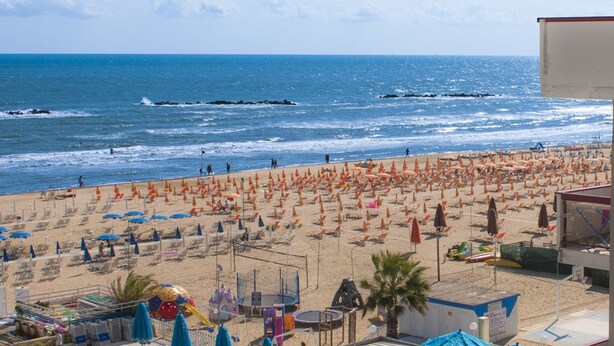 Una playa con numerosos sombrillas y tumbonas, un cartel que dice "HOTEL PALMAR" y vistas al océano.