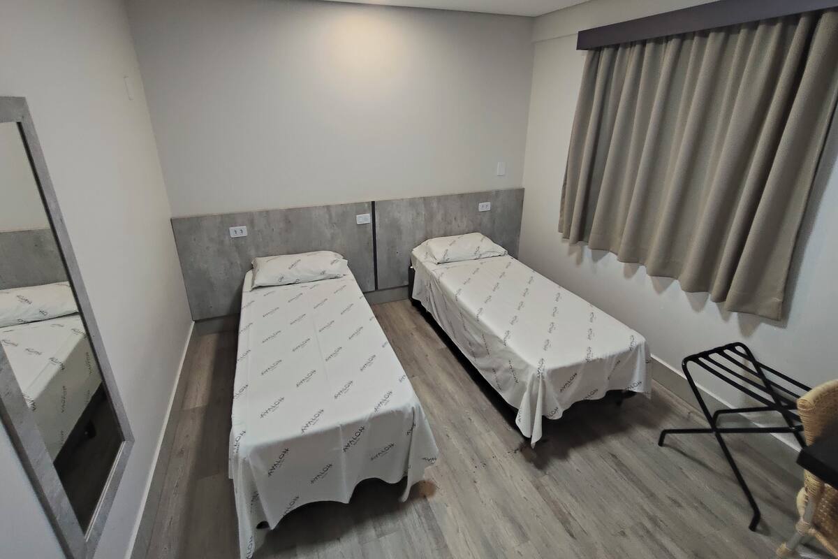 A small, clean room with two single beds, a window with curtains, a chair, and a mirror.
