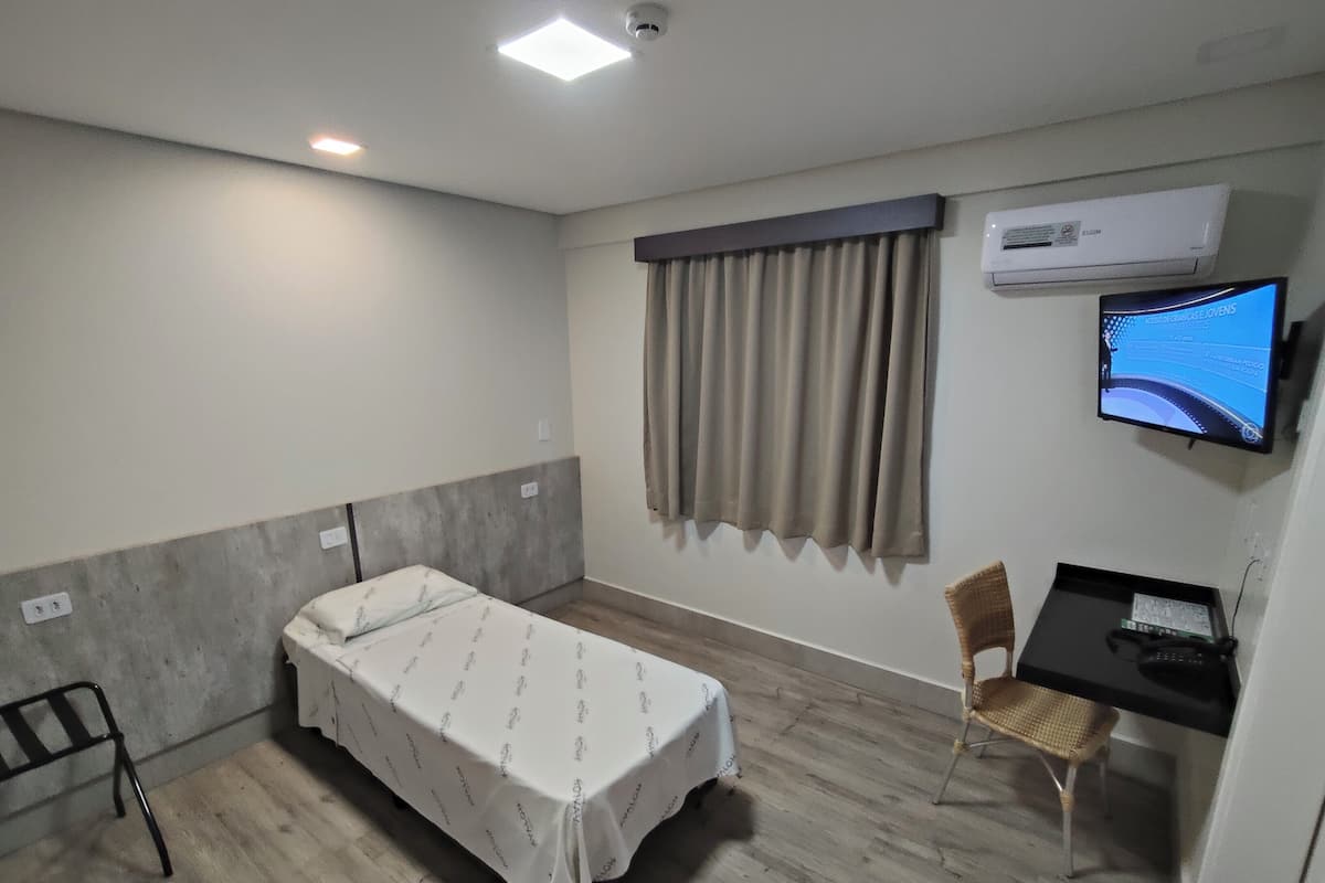 A hotel room with a single bed, a desk with a chair, a television, and an air conditioning unit.