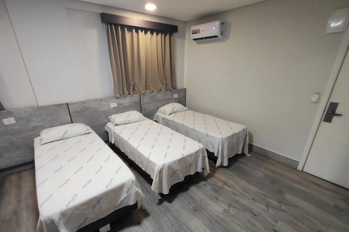 A room with three single beds, a window with curtains, an air conditioning unit, and a door.