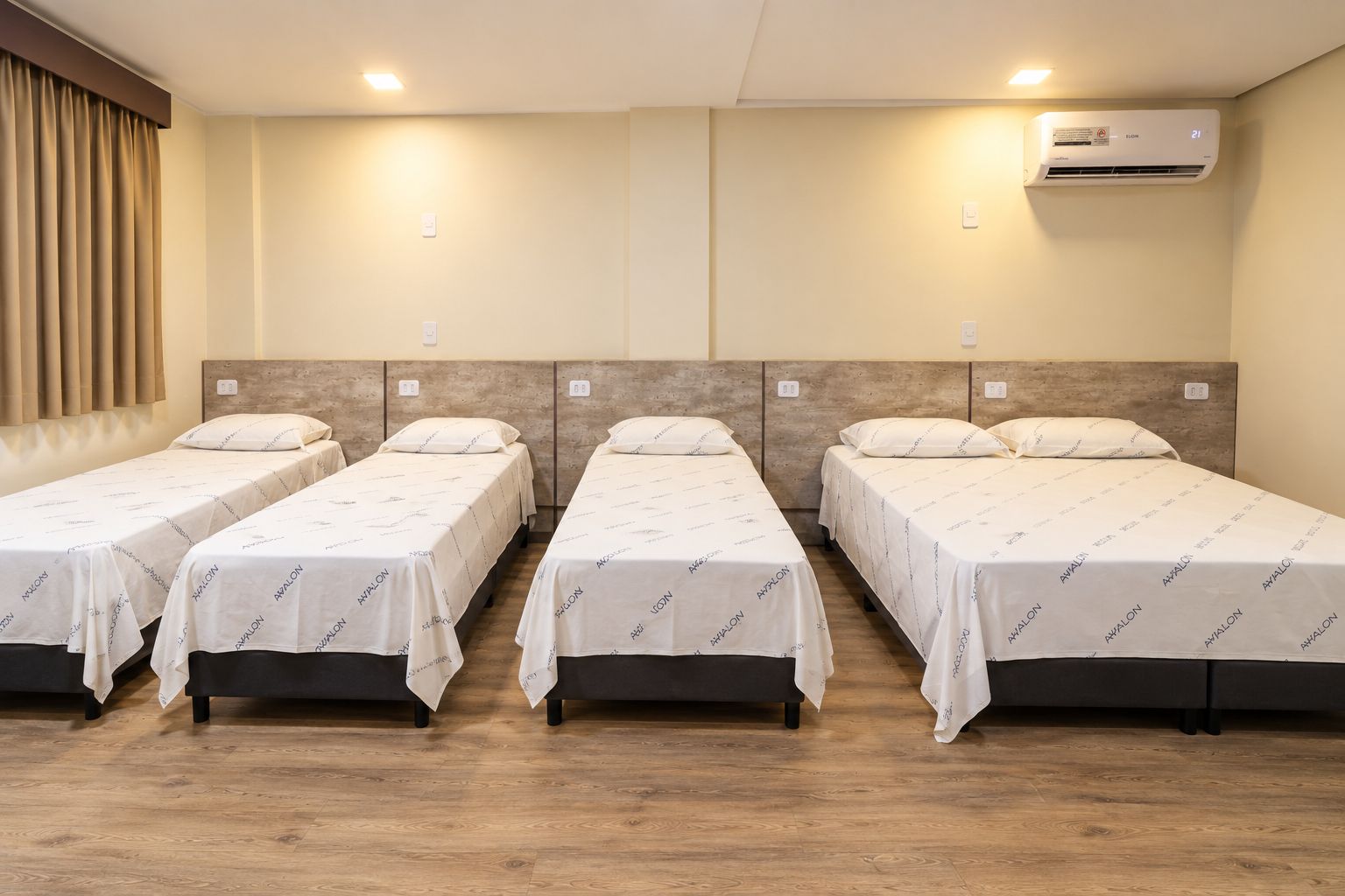 A hotel room with four single beds, a wooden floor, a wall-mounted air conditioner, and a window with curtains.