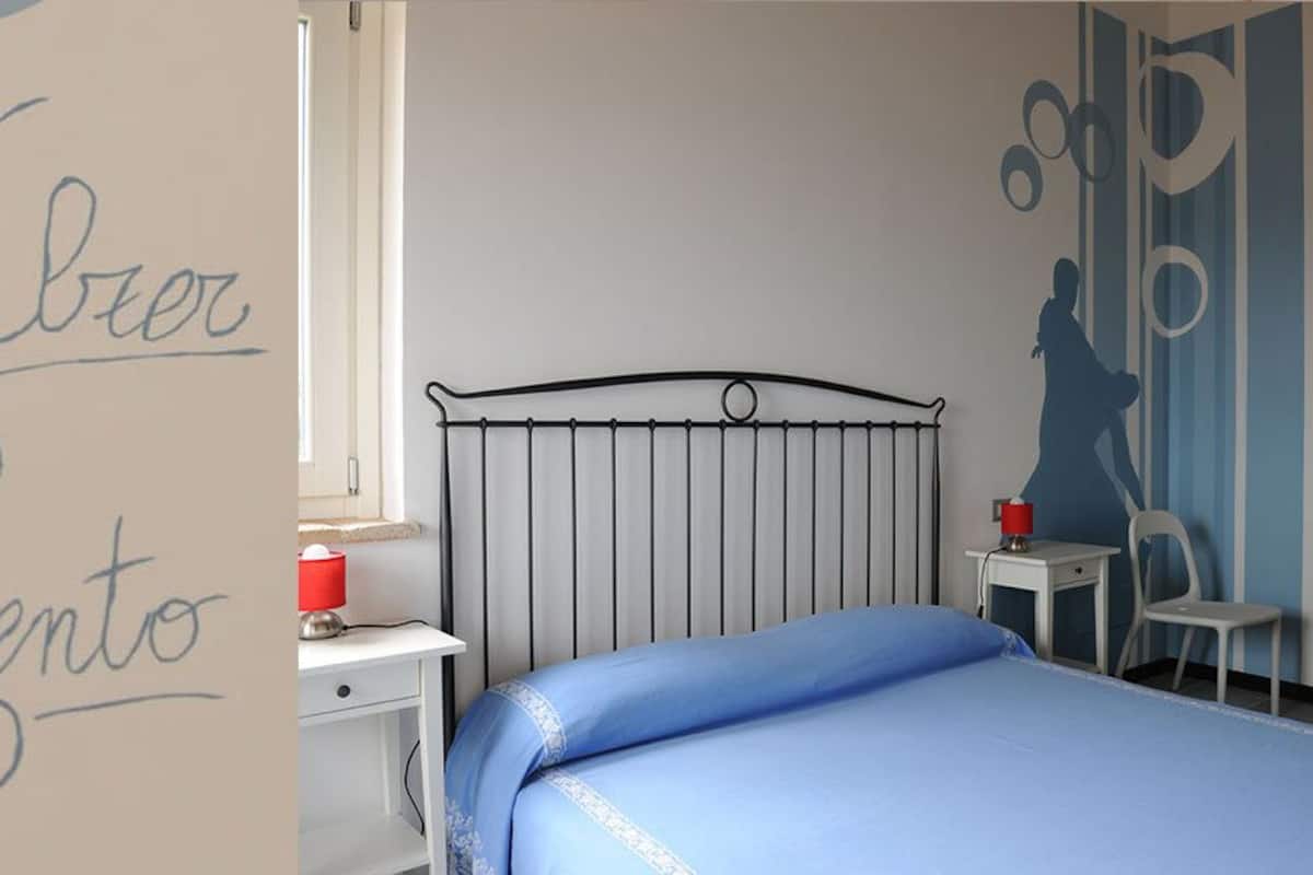A bedroom with a metal bed frame, a blue bedspread, a bedside table with a red lamp, and a small chair.