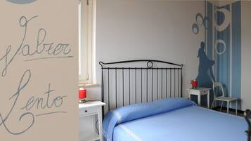 A bedroom with a metal bed frame, a blue bedspread, a bedside table with a red lamp, and a small chair.