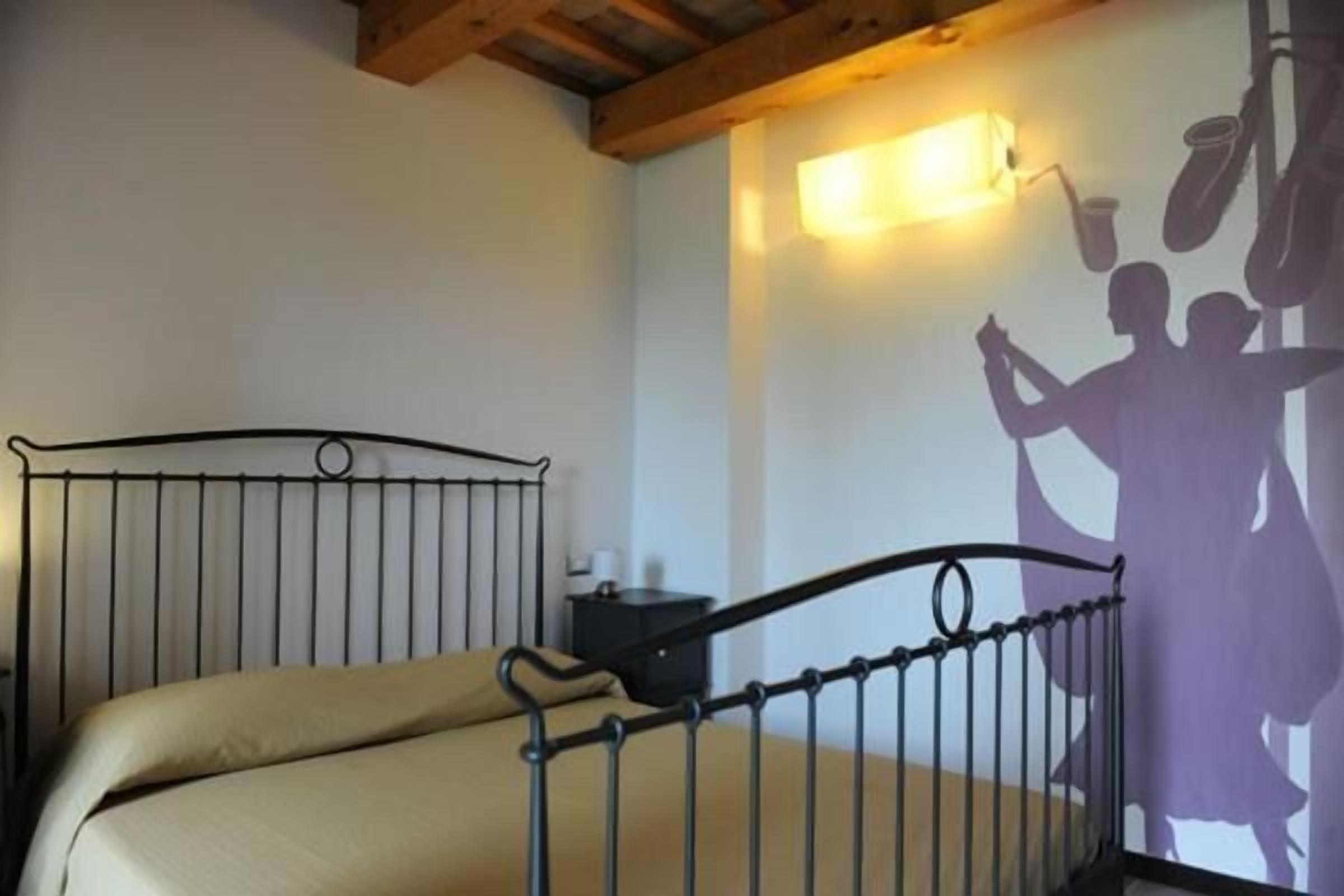Un dormitorio con estructura de cama metálica, colchón color beige, mesita de noche e iluminación fijada a la pared.