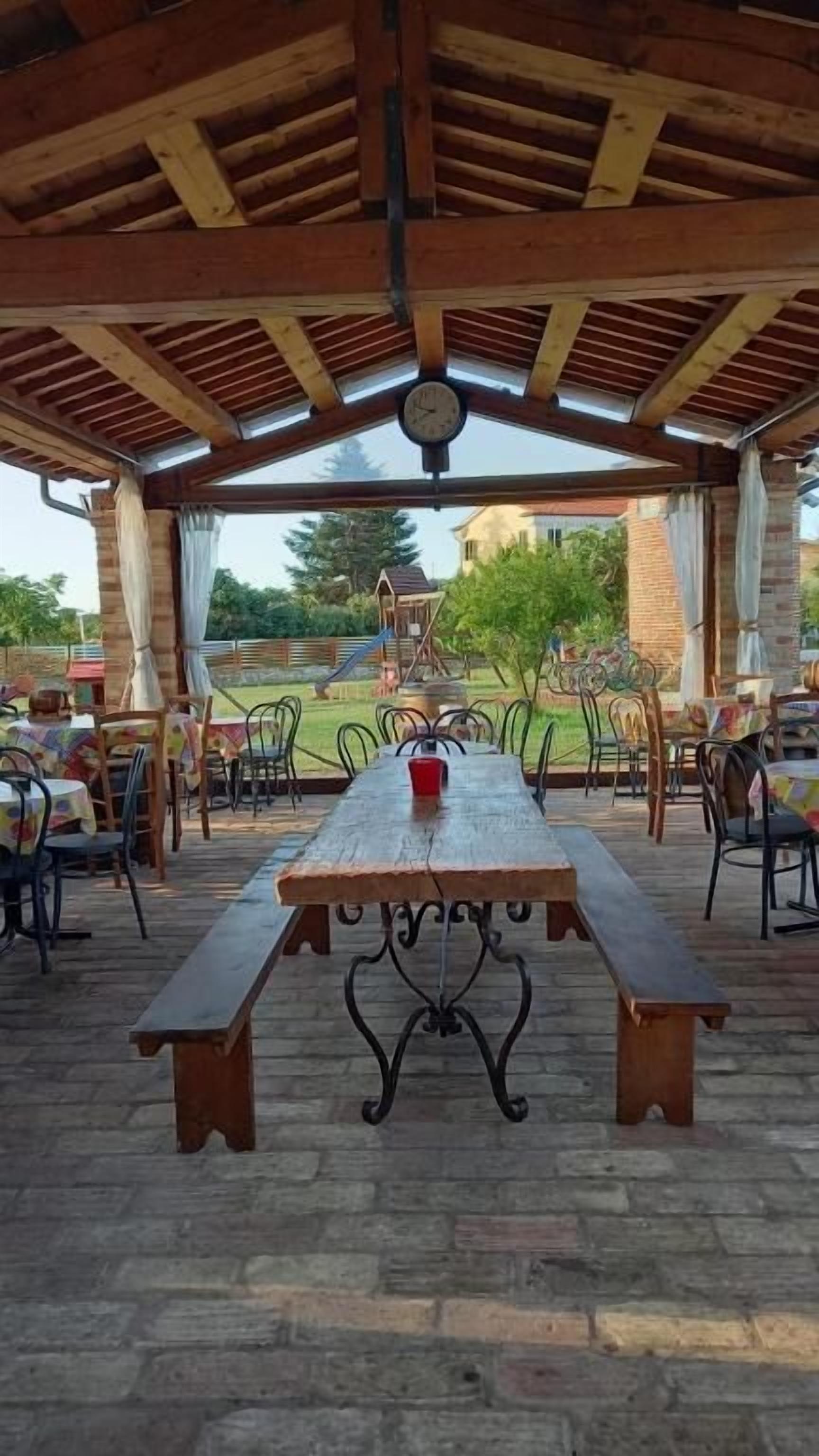 An outdoor covered patio with a wooden table and benches, surrounded by chairs.