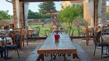 An outdoor covered patio with a wooden table and benches, surrounded by chairs.