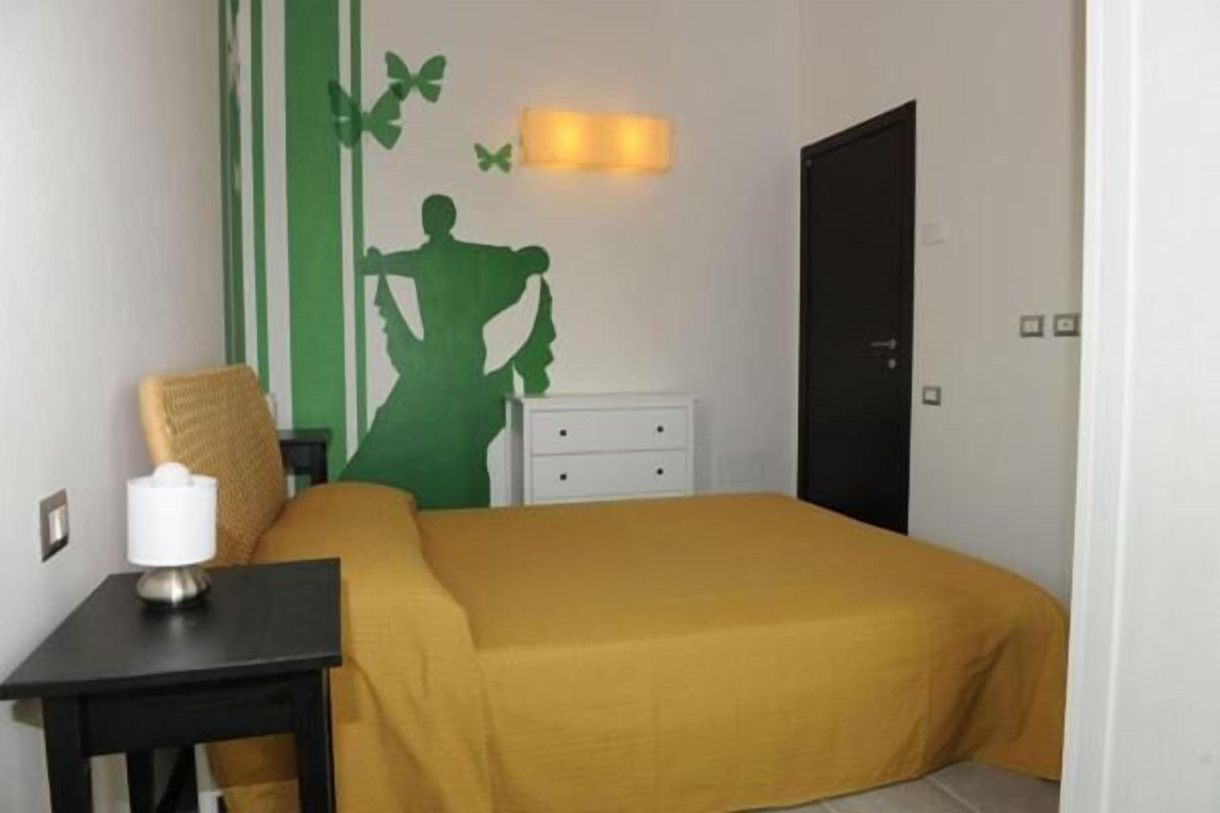 Una habitación con cama, mesita de noche, lámpara, silla, pared con silueta verde y una puerta.