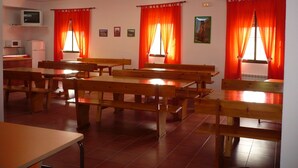 Un comedor con bancos y mesas de madera, una zona de cocina con microondas y refrigerador, y ventanas con cortinas anaranjadas.