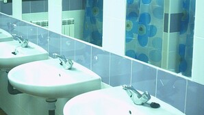 Baño público con tres lavamanos, un espejo y zona de ducha con cortinas azules estampadas con motivos florales.
