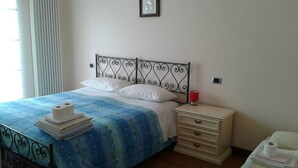 A neatly made bed with a blue floral bedspread, white pillows, and a stack of white towels on the side. There is a bedside table with a red lamp and a drawer. - B&B Paradiso (Loreto)