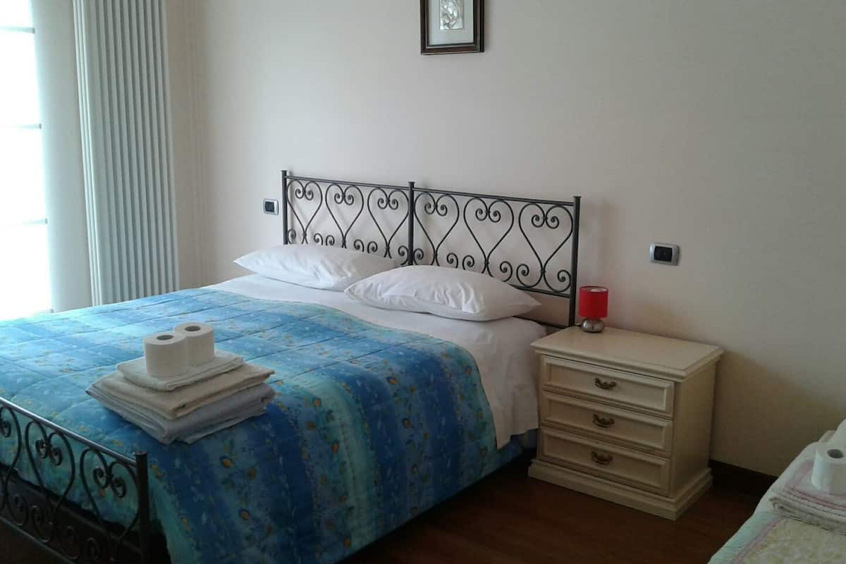 Cama bem arrumada com colcha floral azul, almofadas brancas e uma pilha de toalhas brancas ao lado. Tem uma mesa de cabeceira com abajur vermelho e uma gaveta.