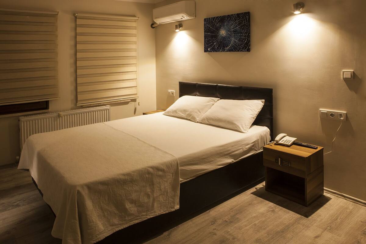 Una cama bien tendida con sábanas blancas en una habitación con suelo de madera, un radiador y aire acondicionado de pared.