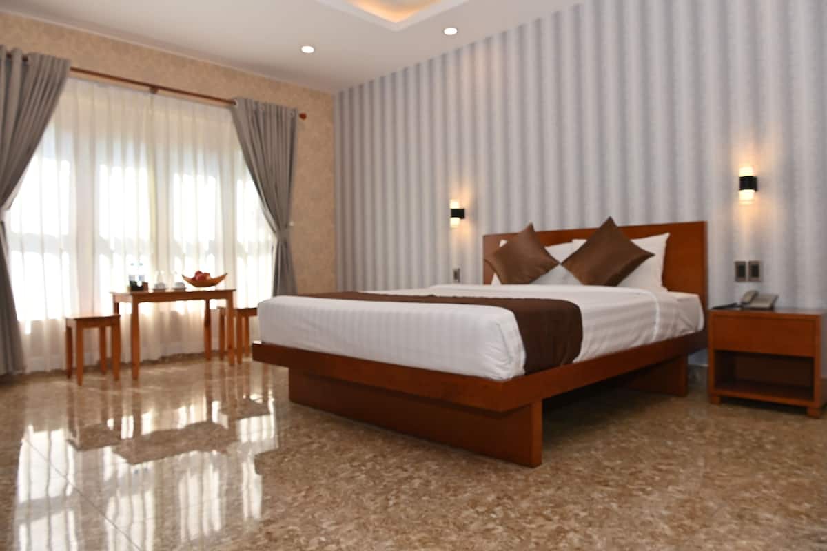 Ein modernes Hotelzimmer mit einem großen Bett, einem Holznachttisch, einem Schreibtisch mit Stuhl und einem großen Fenster mit Vorhängen.