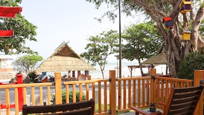 Une terrasse en bois avec des bancs donnant sur une plage, une structure à toit de chaume et des nichoirs colorés accrochés à un arbre.