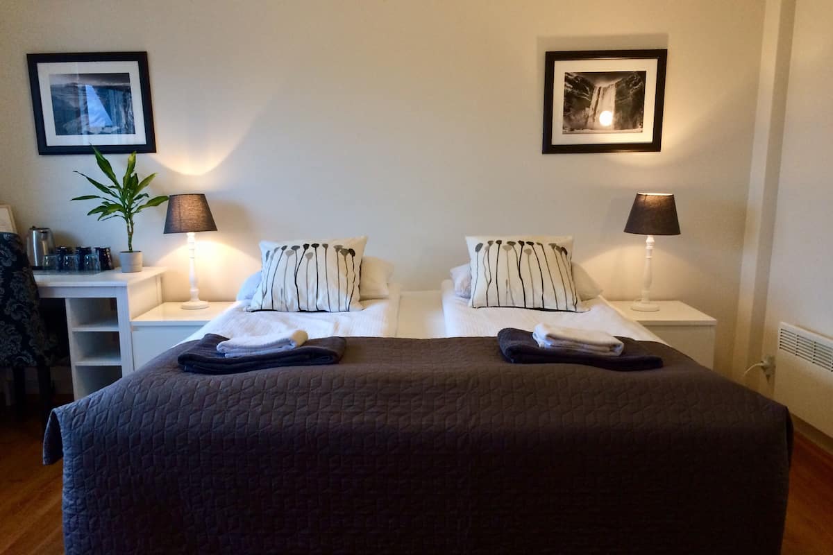 Un lit double avec une couverture sombre, deux tables de chevet avec des lampes, une plante en pot et deux tableaux encadrés accrochés au mur.