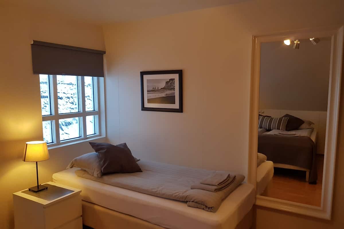 Une chambre à coucher avec un lit, une table de chevet avec une lampe, un tableau encadré au mur et une fenêtre donnant sur des arbres.