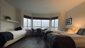 A corner room with two beds, a desk, and a chair. There are large windows providing a view of the outside.