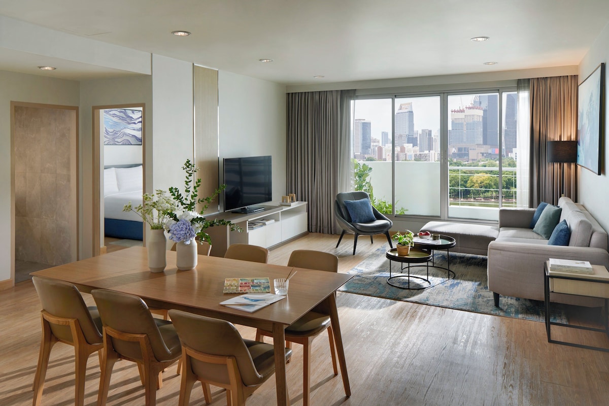 Quarto de hotel moderno com área de refeições, sala de estar com sofá e poltronas, televisão e vista para a cidade.
